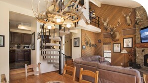 A spacious living area with a brown leather sofa, wooden dining table, and a chandelier. A staircase leads to a loft bedroom.