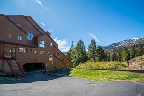 A large, multi-story building with a garage entrance, surrounded by a well-maintained lawn and trees, with mountains in the background.