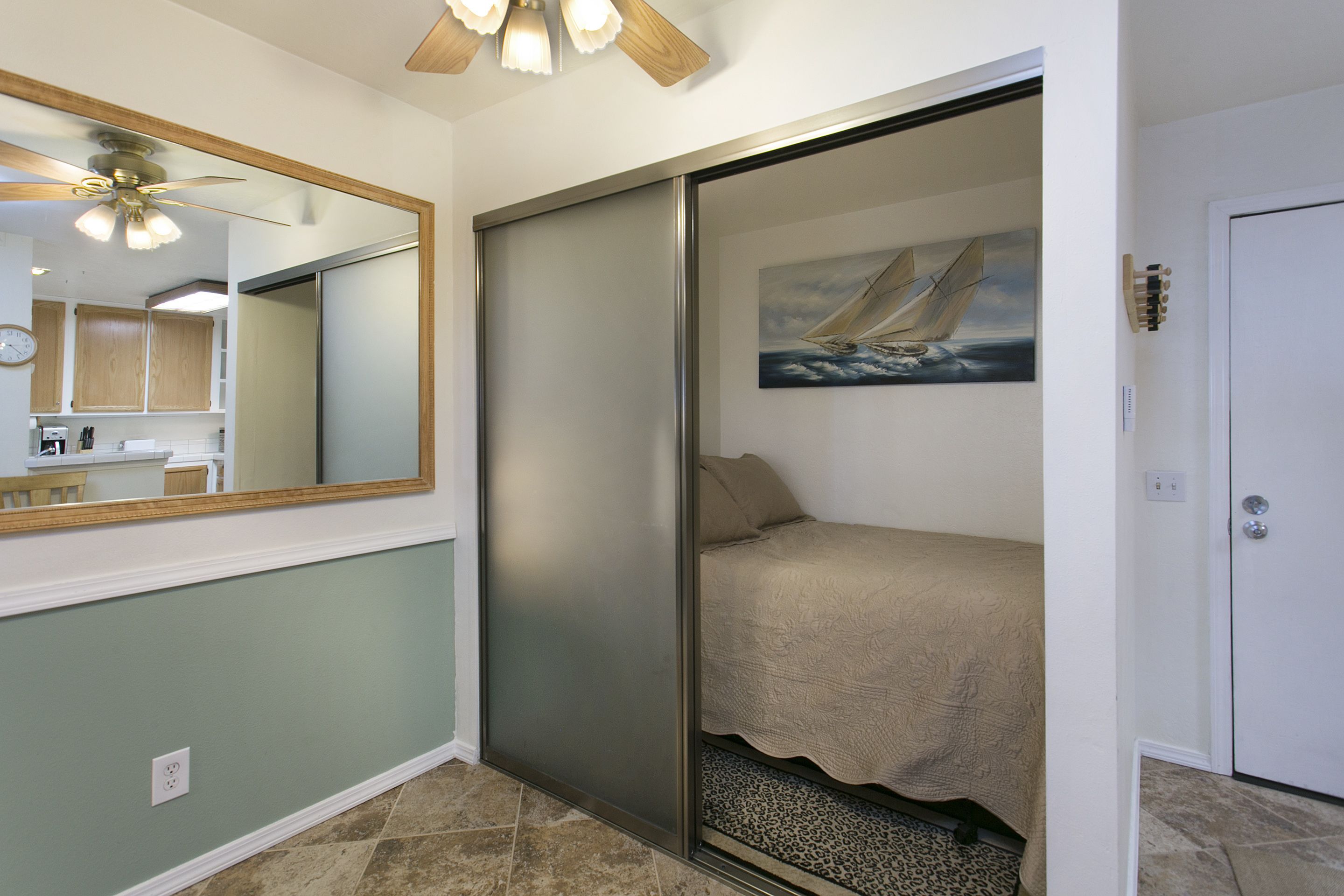 A bedroom with a sliding door, a bed, a painting of a sailboat, a ceiling fan, and a mirror.