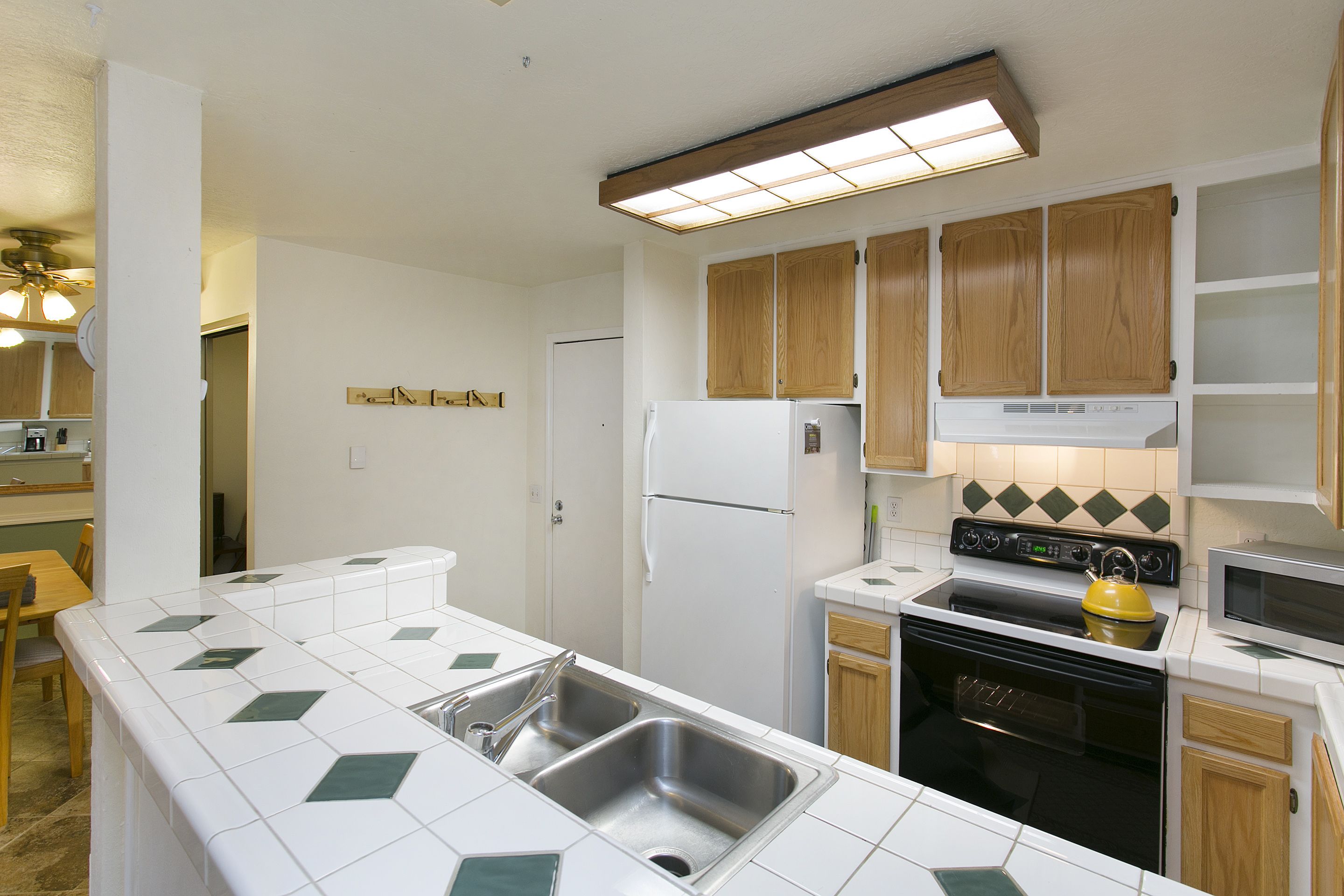 A kitchen with white tiles, wooden cabinets, a refrigerator, stove, microwave, and a sink.