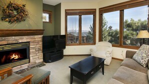 A living room with a stone fireplace, a flat-screen TV, a sofa, a coffee table, and large windows offering a view of trees and mountains. - Eagle Run by 101 Great Escapes (Mammoth Lakes)