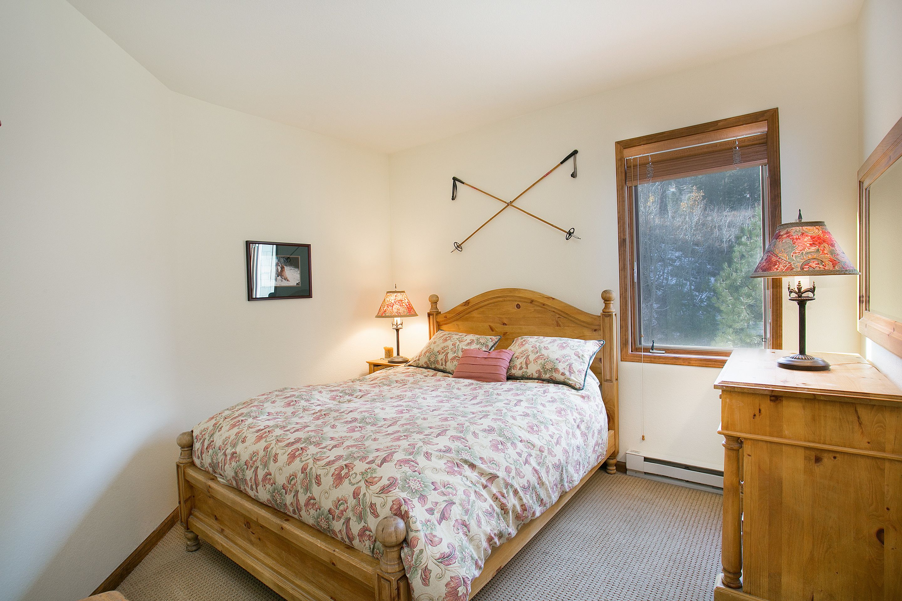 A bedroom with a wooden bed, a nightstand with lamps, a window with a view of trees, and a wooden dresser.