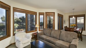 A living room with a sofa, armchair, and a table with chairs. There are large windows with blinds, a lamp, and a vase with sunflowers. - Eagle Run by 101 Great Escapes (Mammoth Lakes)