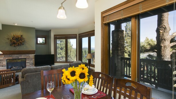 A dining area with a table set for two, a fireplace, and large windows offering a view of trees and a mountain. - Eagle Run by 101 Great Escapes (Mammoth Lakes)