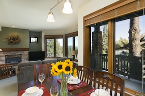 Un coin repas avec une table dressée pour deux personnes, une cheminée et de grandes fenêtres donnant sur des arbres et une montagne.