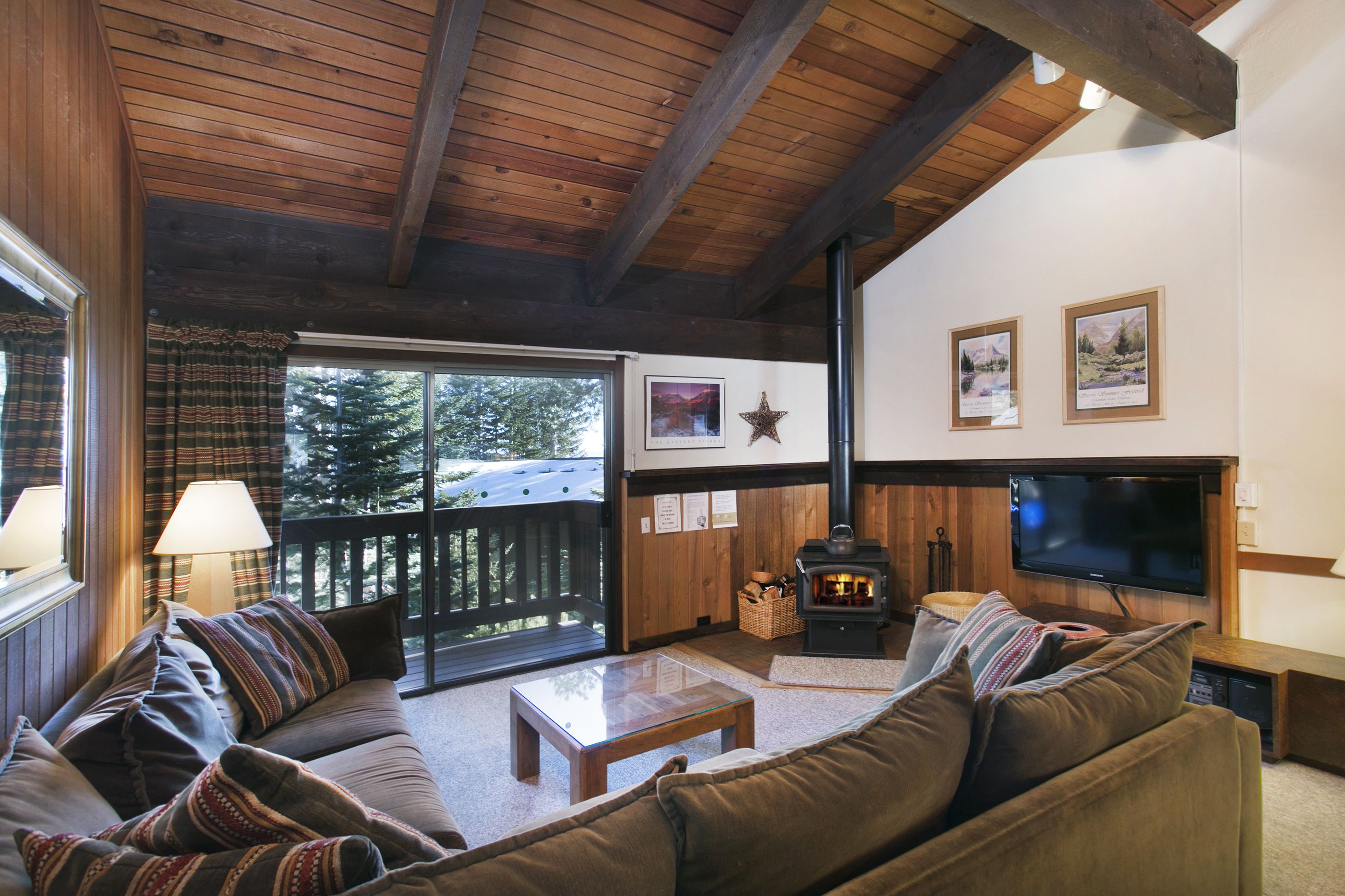A cozy living room with a wooden ceiling, a fireplace, a television, and a large window overlooking a deck.