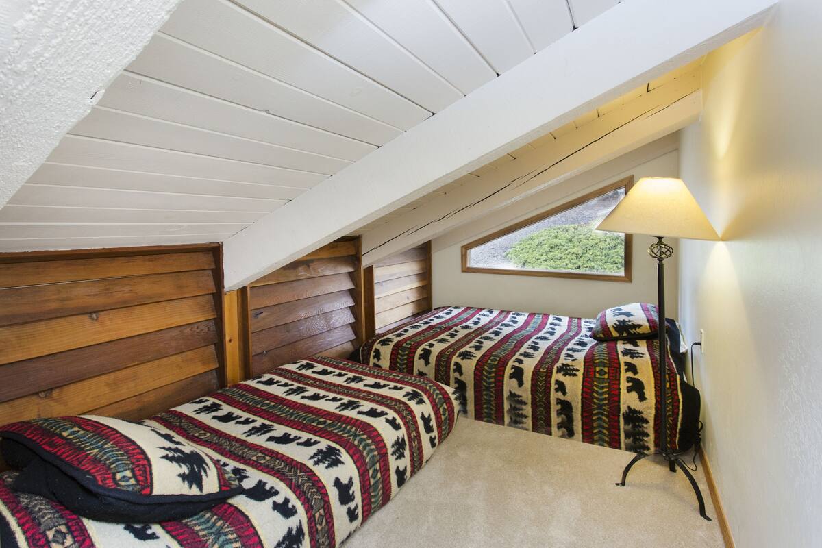 A small, cozy attic bedroom with two beds, a window, and a lamp.