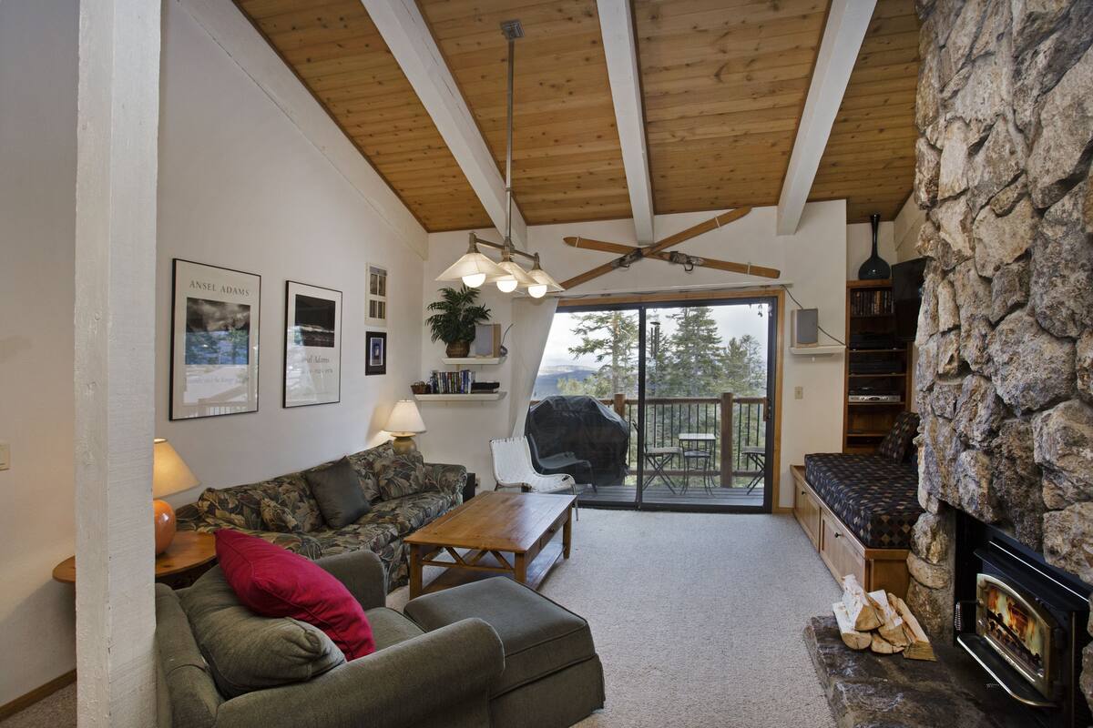A cozy living room with a fireplace, a ceiling fan, and a large window overlooking a deck.