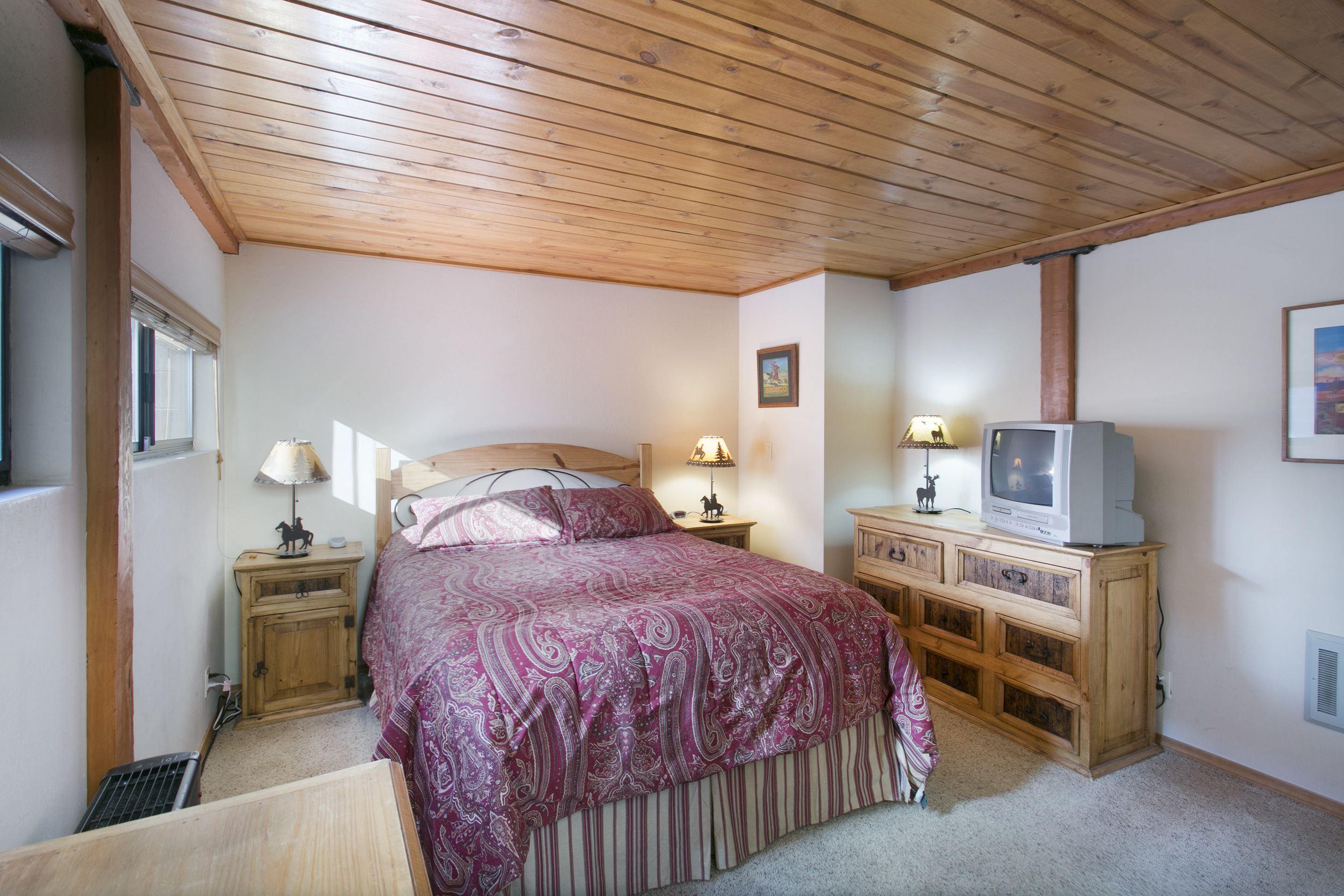 A bedroom with a wooden ceiling, a bed with a patterned bedspread, two bedside lamps, a television on a wooden cabinet, and a window with blinds.