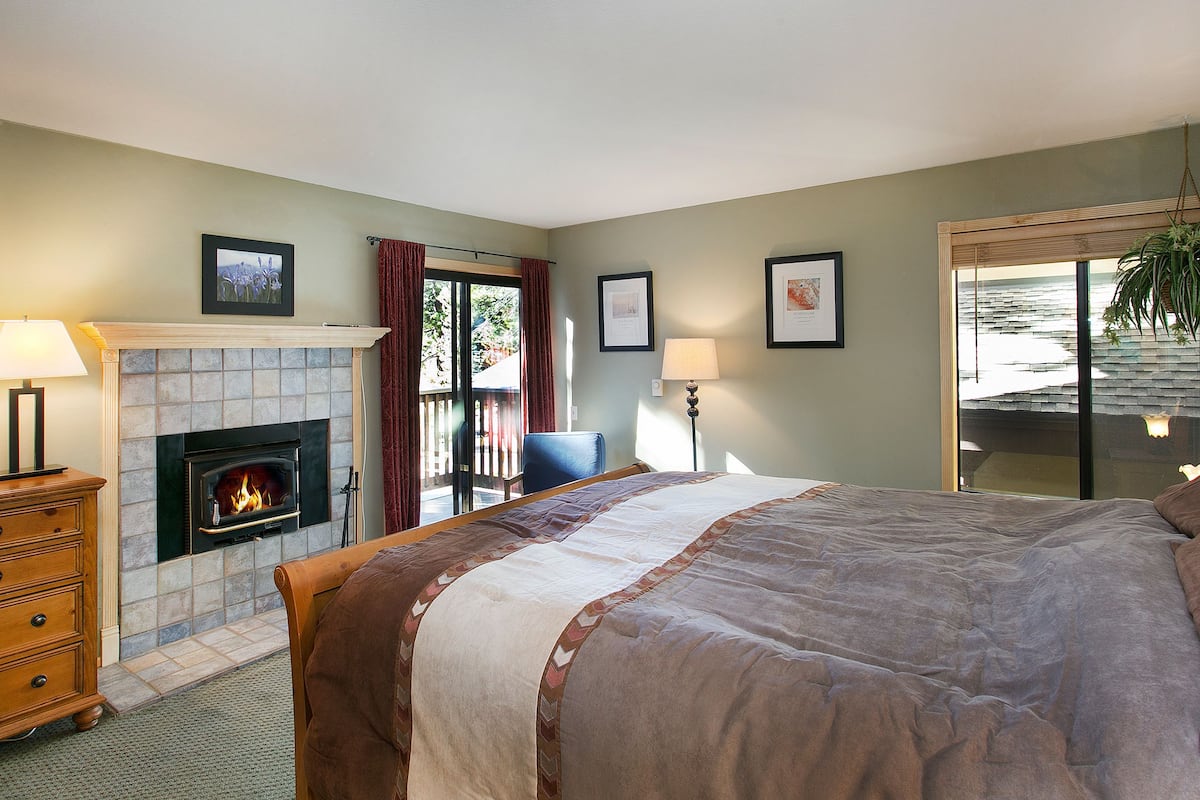 Une chambre avec une cheminée, un lit recouvert d’une couverture marron, une chaise et une vue à travers une porte vitrée.