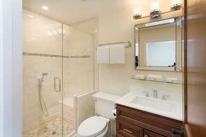 Un baño moderno con ducha envuelta en vidrio, inodoro blanco, tocador de madera con espejo y estante de vidrio sobre el lavabo.