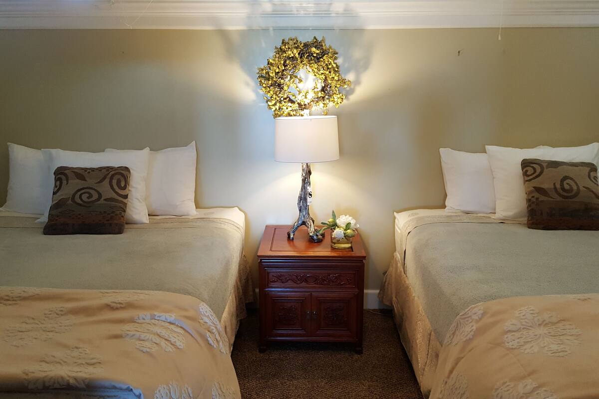 A hotel room with two beds, each with a patterned pillow, a nightstand with a lamp and a vase of flowers, and a decorative wall piece above the beds.