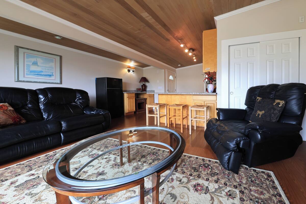 A living room with a black leather sofa, a glass-top coffee table, a black leather recliner, and a wooden ceiling.