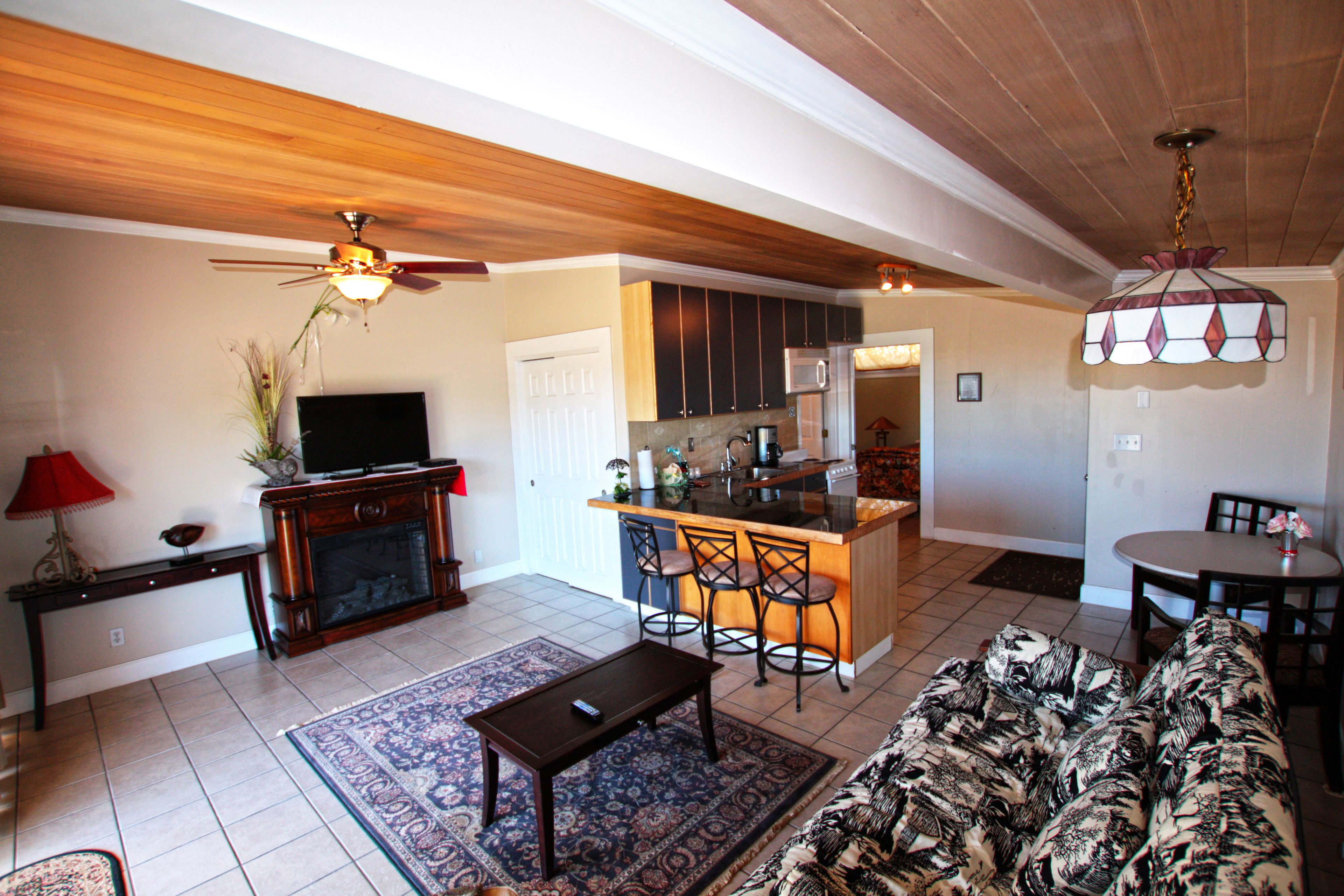A spacious living area with a sofa, coffee table, and a TV. A kitchen with a bar and seating is visible in the background.