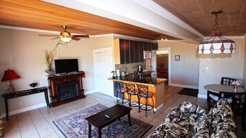 A spacious living area with a sofa, coffee table, and a TV. A kitchen with a bar and seating is visible in the background.