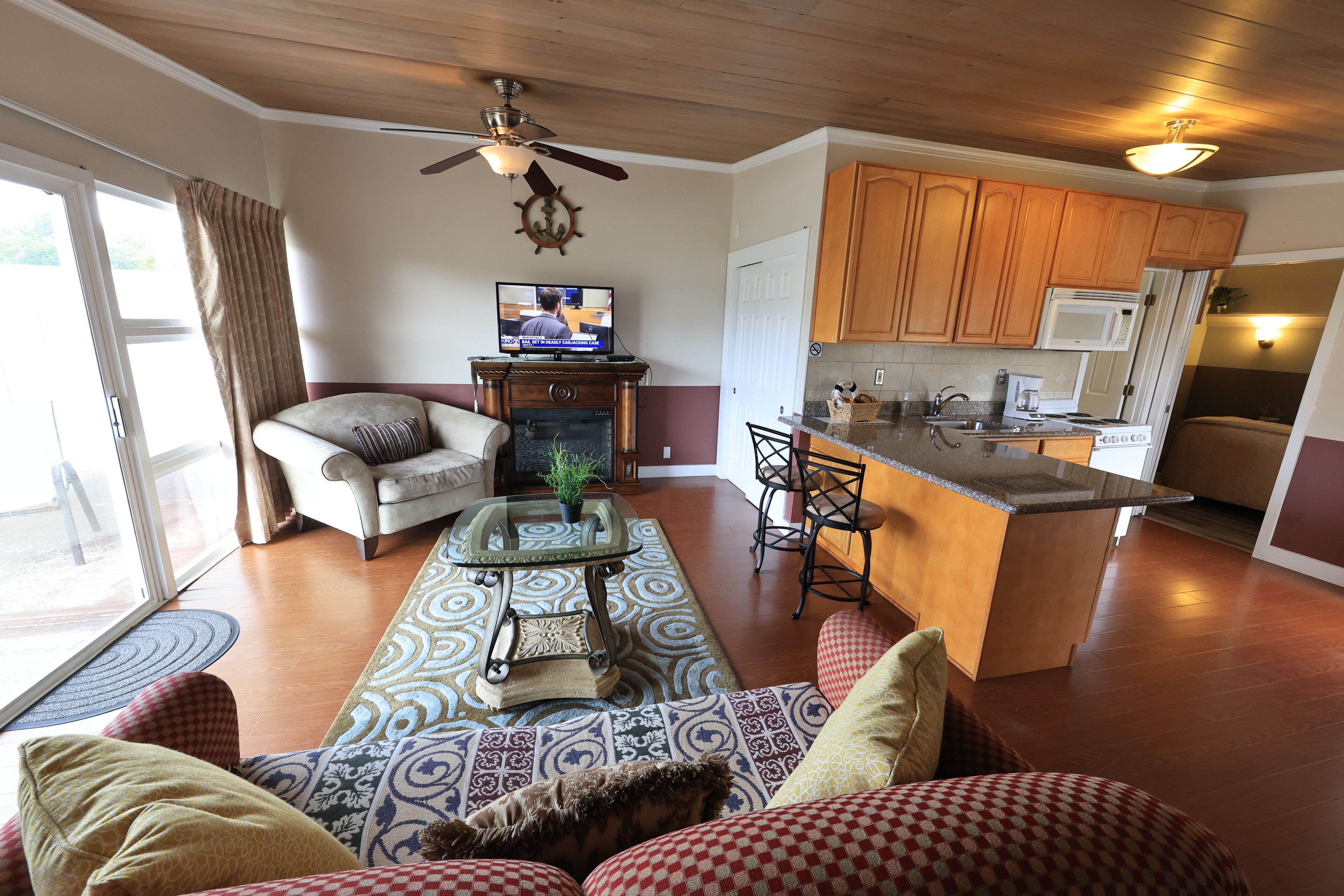 A living room with a sofa, a coffee table, and a television. There is a kitchen area with wooden cabinets and a microwave.