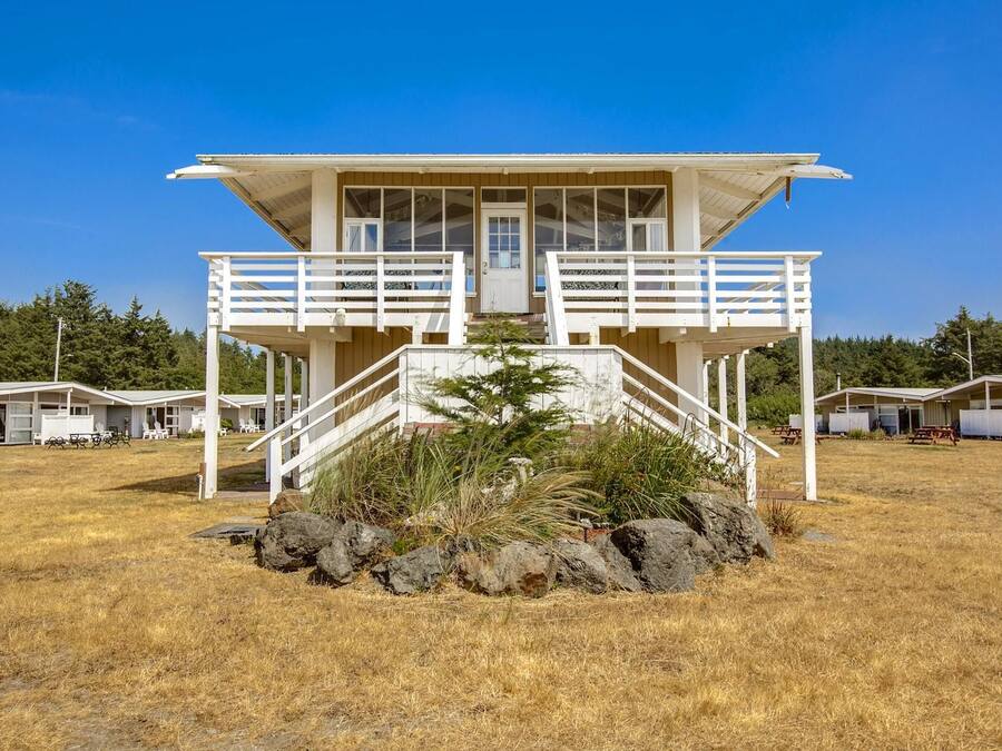 A two-story beach house with a balcony, a front porch, and a small garden area with rocks.