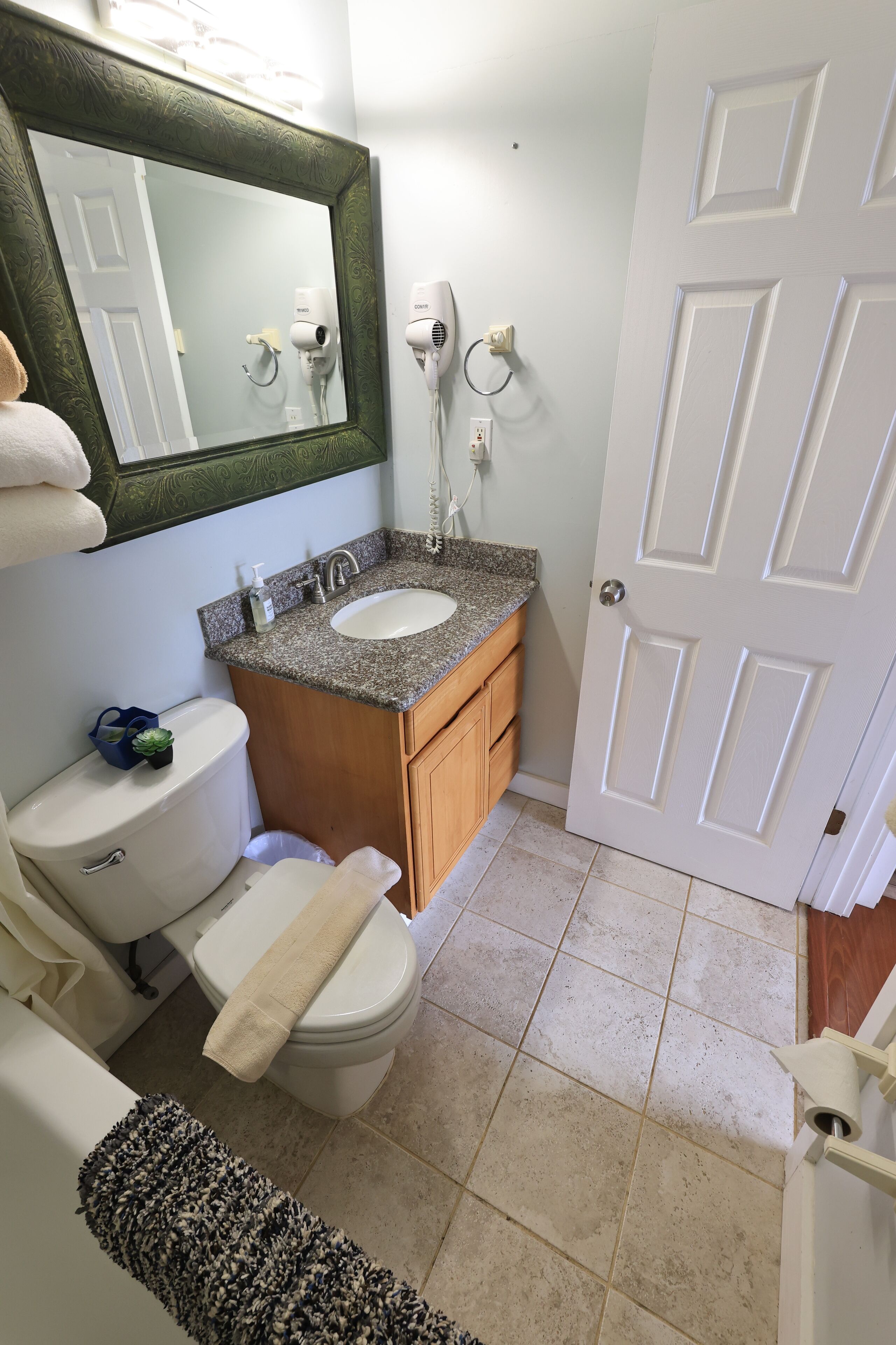 A bathroom with a white toilet, a sink with a granite countertop, a wooden cabinet, a mirror with an ornate frame, and a white door.