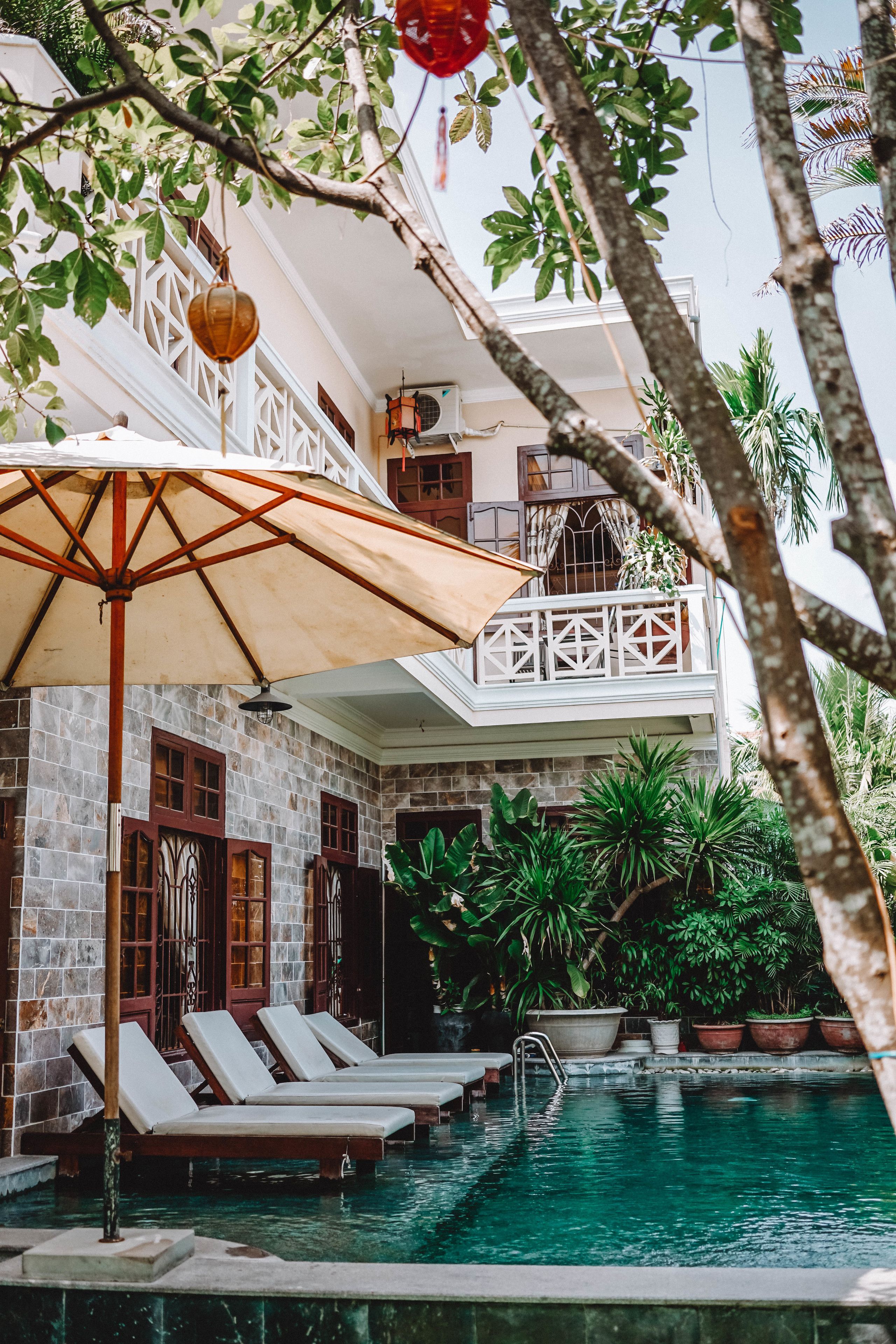 A pool area with lounge chairs and an umbrella, surrounded by lush greenery and a two-story building with balconies.