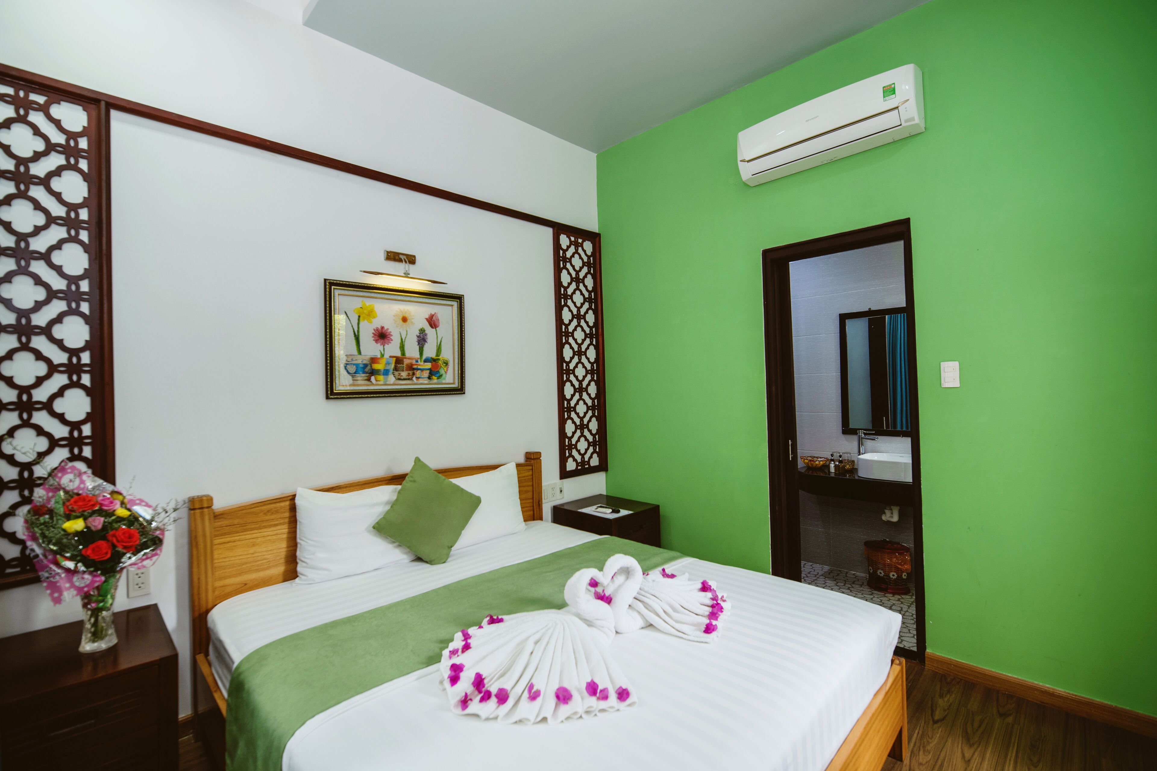 A hotel room with a green wall, a wooden bed, a green throw blanket, a bouquet of flowers, and a framed picture on the wall.