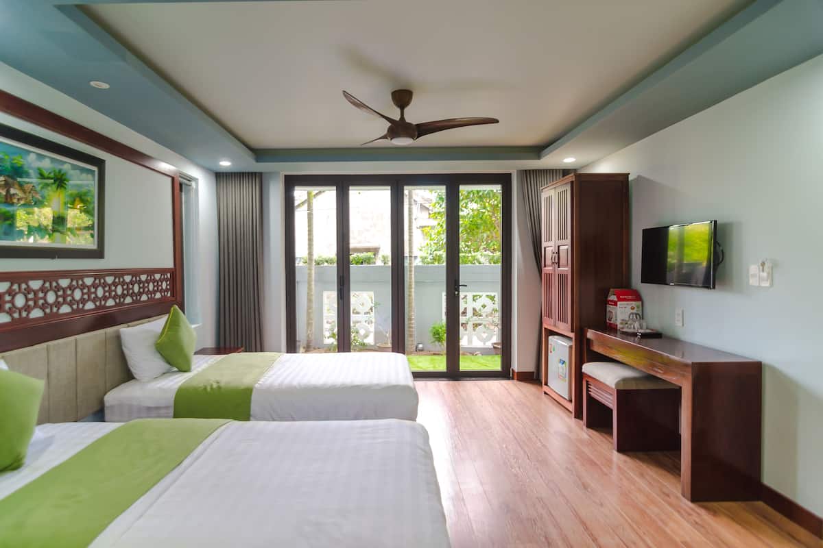 A hotel room with two beds, a desk, a television, and a balcony with a view of greenery.