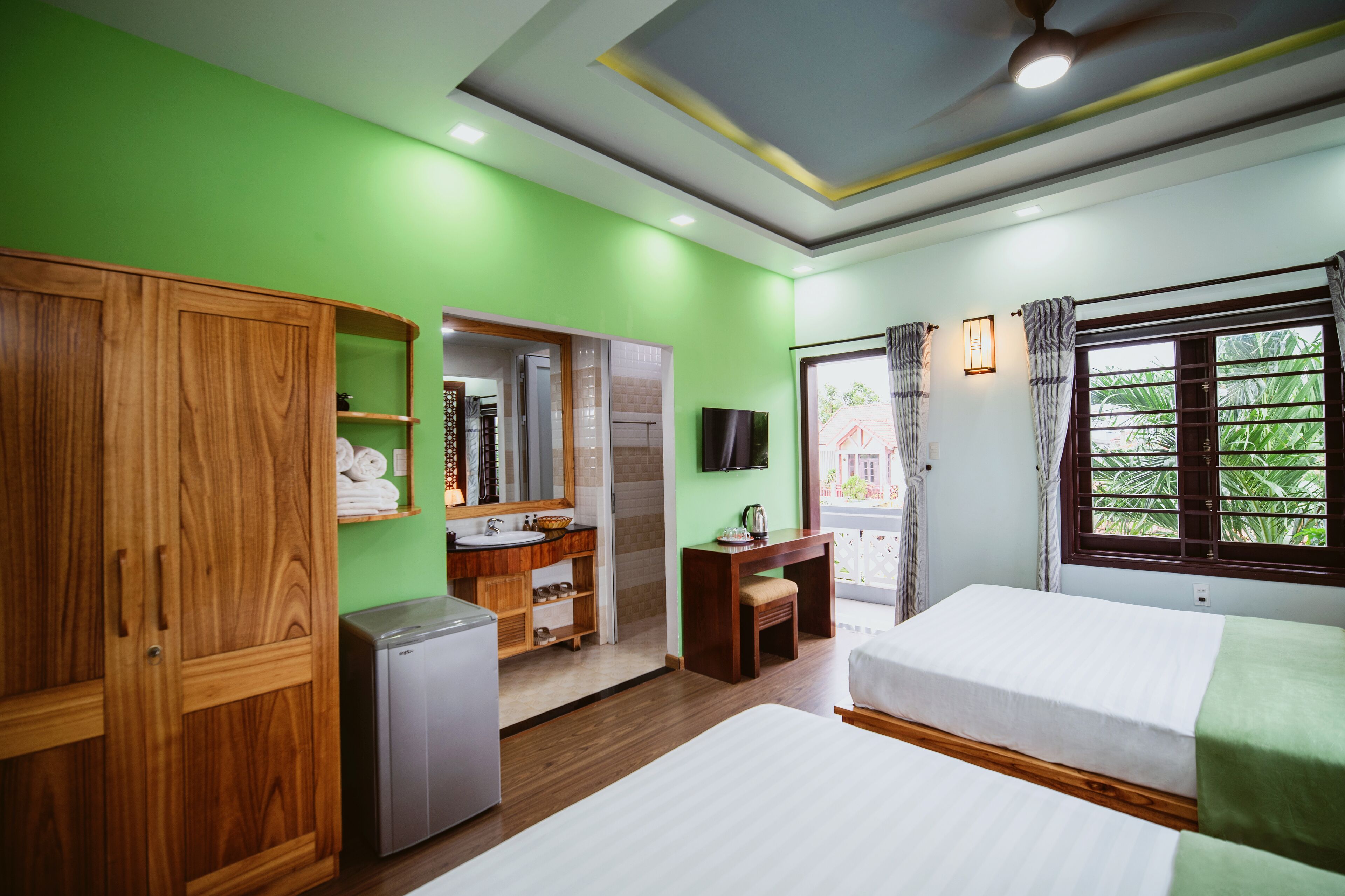A hotel room with a green wall, wooden wardrobe, a small desk, a bed, and a window with curtains.