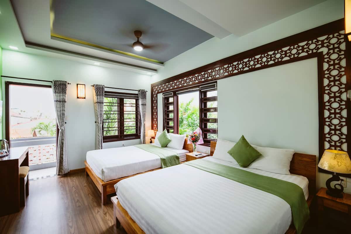 A hotel room with two beds, wooden flooring, a ceiling fan, and large windows with curtains.