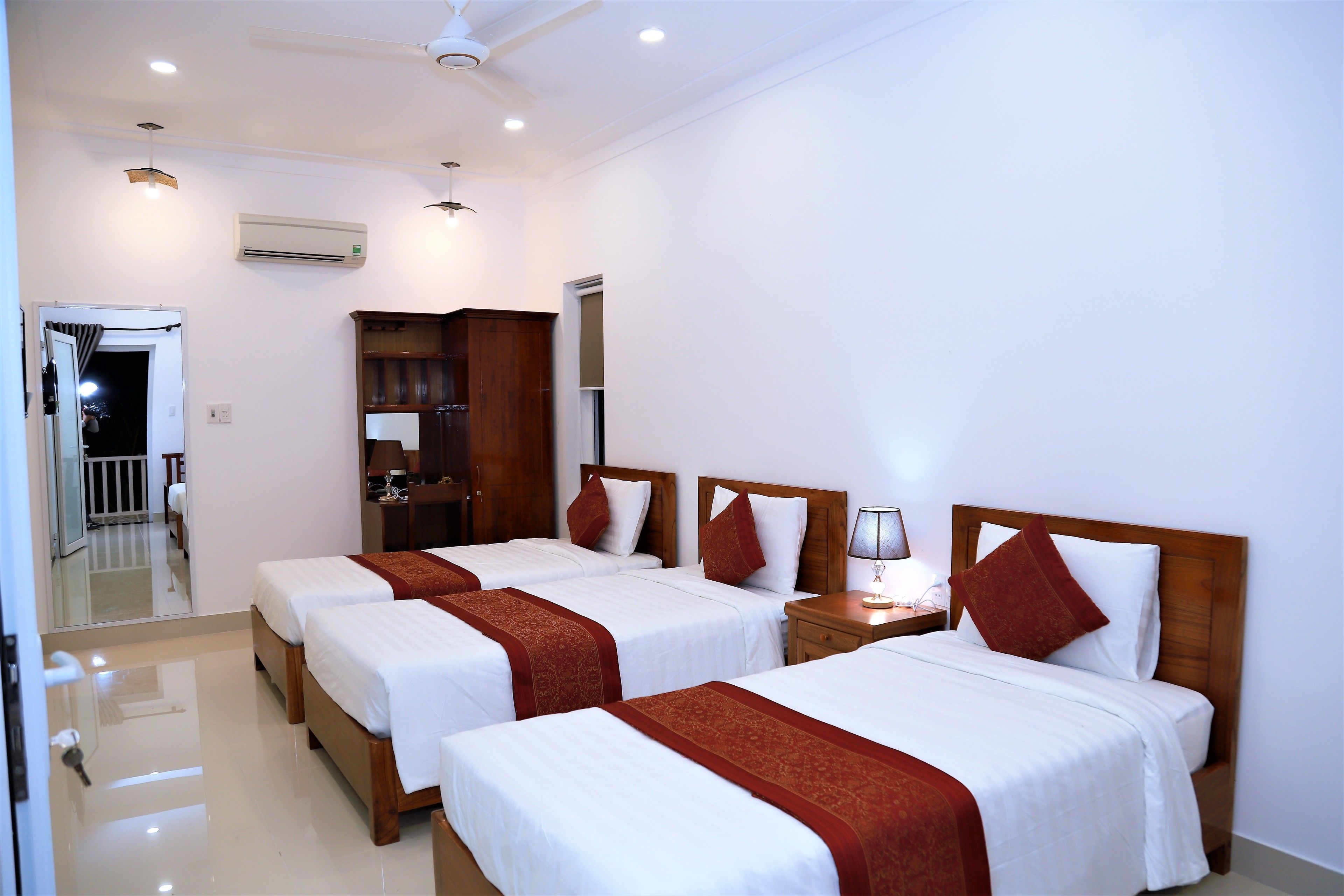 A hotel room with three beds, a wooden wardrobe, a mirror, and a ceiling fan.
