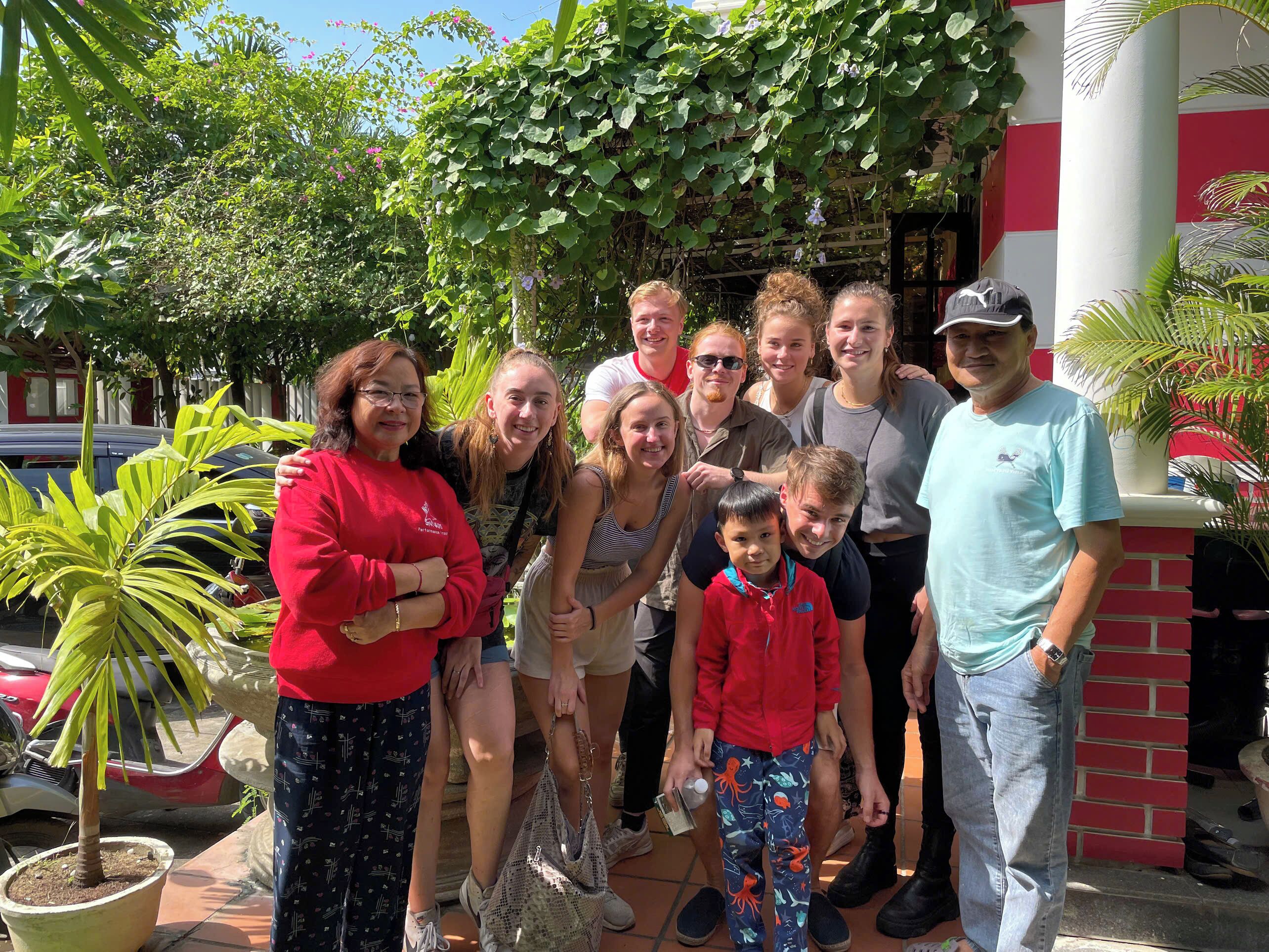 A group of people posing in front of a building with a red and white facade, surrounded by greenery and potted plants.