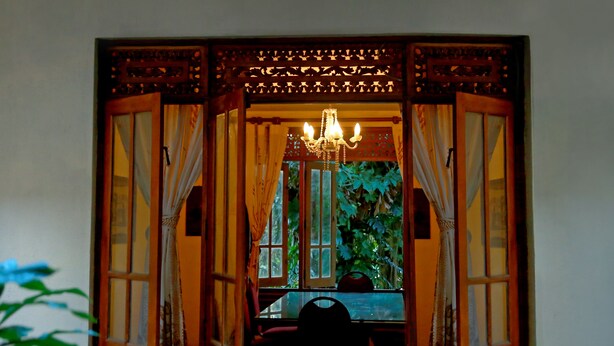 Una ventana abierta con cortinas, una araña y vistas al verdor exterior.