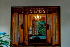 Una ventana abierta con cortinas, una araña y vistas al verdor exterior.