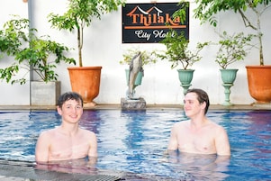 Piscina con dos personas, una estatua de delfín y un letrero del Hotel Thilaka City.