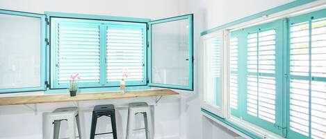 A room with a wooden desk, two metal stools, a small potted plant, and a window with white shutters.