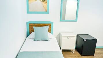 Una cama individual con cabecera de madera, una mesita de noche con cajón, un refrigerador negro pequeño, un cuadro enmarcado de un faro y un espejo en la pared.