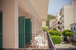 Un balcón con sillas de plástico blanco, un ventanal con persiana verde y vistas a una calle estrecha con edificios y vegetación.