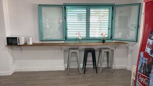 A kitchenette with a microwave, kettle, and a window with blinds. There are two stools and a Coca-Cola poster. - Hostal Sunset Ibiza (Sant Antoni de Portmany)