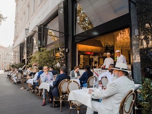An outdoor café with tables and chairs, a chef in uniform, and a building with a sign. - Hotel Grand Ferdinand Vienna (Vienna)