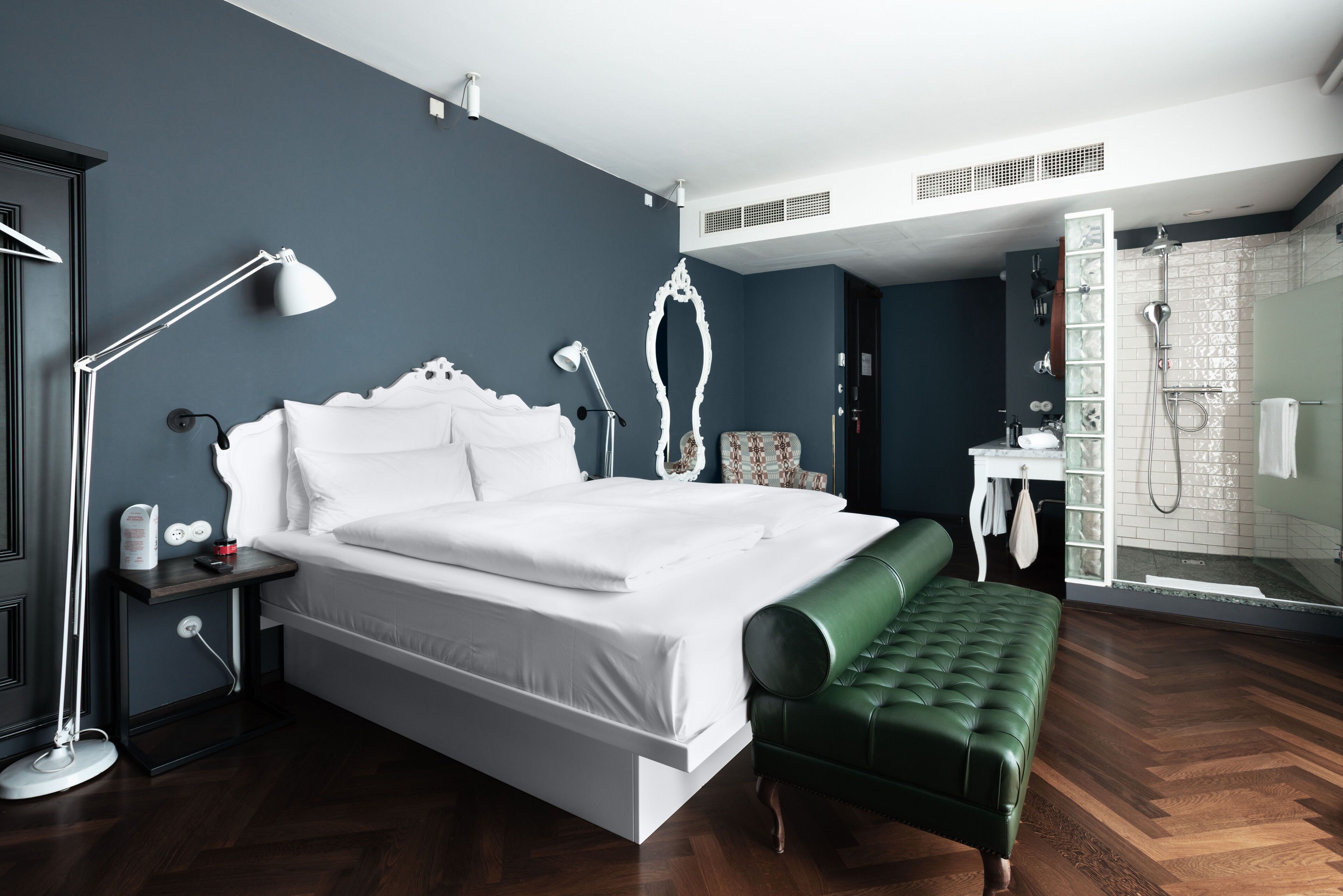 A modern hotel room with a large bed, a green bench, a bedside table with a lamp, and a bathroom with a shower.