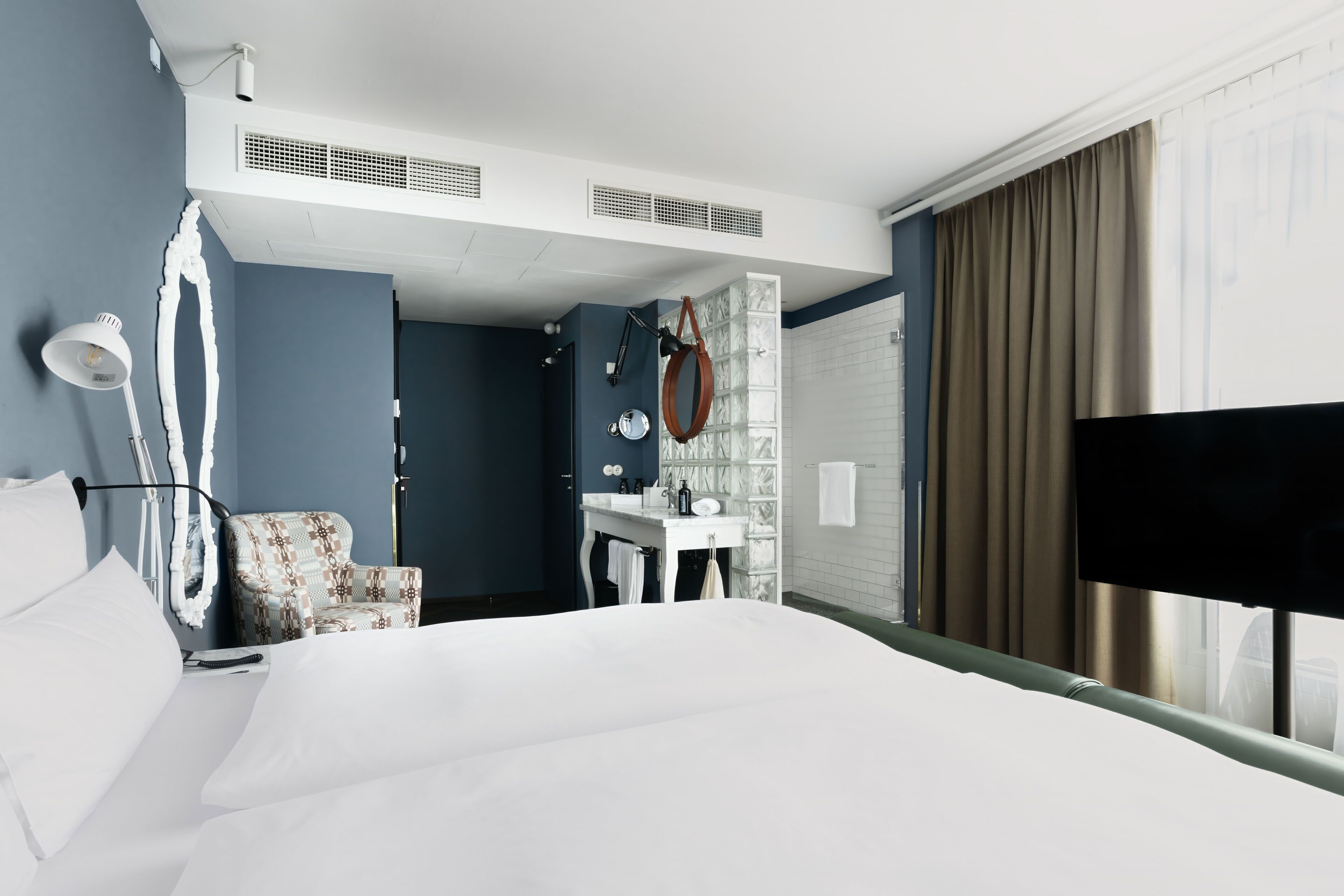 A hotel room with a bed, a chair, a desk, a mirror, a window with curtains, and a TV.
