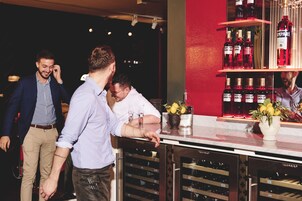Uma área de bar com um refrigerador de vinho, garrafas de bebidas e um vaso com flores.