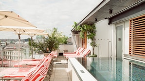 A rooftop pool area with red and white striped lounge chairs, a glass-enclosed pool, and a cityscape view in the background. - Hotel Grand Ferdinand Vienna (Vienna)