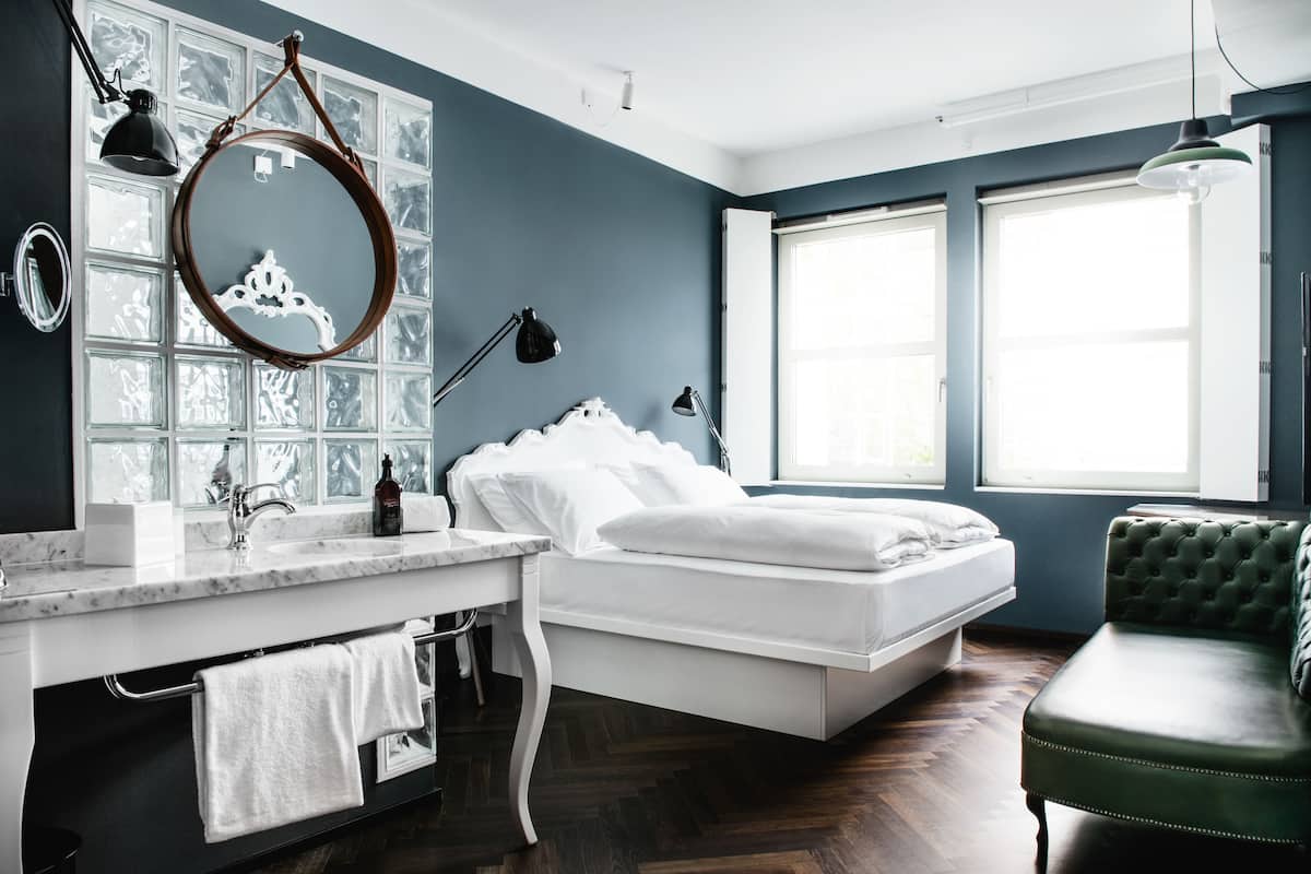 Une chambre à coucher avec un lit, une coiffeuse avec un miroir, un fauteuil vert et de grandes fenêtres.