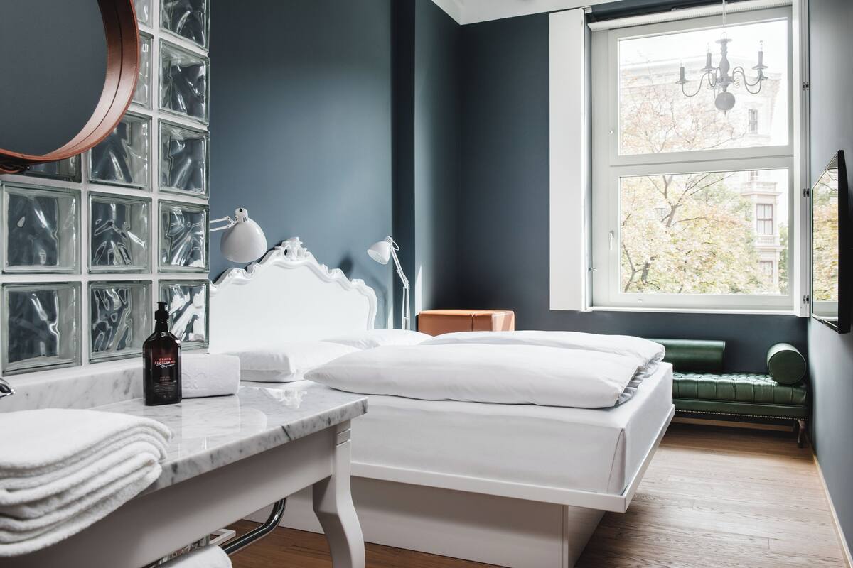 Une chambre avec un lit, une coiffeuse avec un miroir, une fenêtre donnant sur des arbres et un banc vert près de la fenêtre.