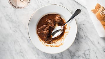 A bowl of stew with a spoon, a glass of wine, and a piece of bread on a marble surface.