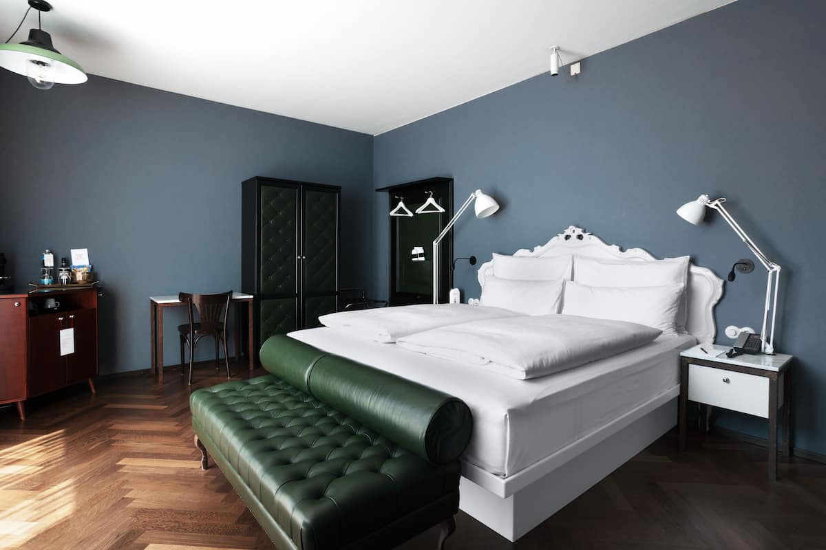 Une chambre à coucher comprenant un lit, un banc recouvert d’un tissu vert, un bureau avec une chaise, une armoire et une table de chevet avec une lampe.