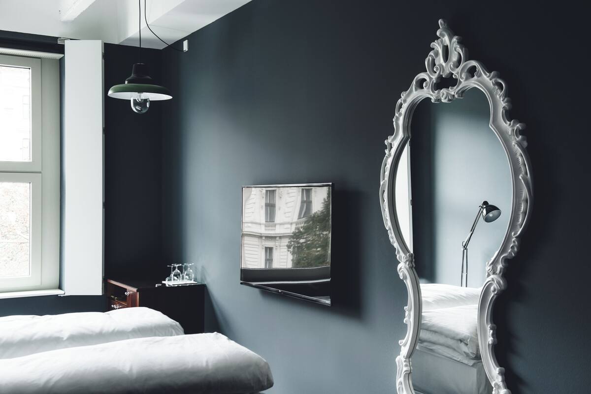 Une chambre à coucher avec un grand miroir décoratif, un lit, une table de chevet avec une lampe et une fenêtre donnant sur un immeuble.
