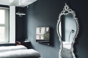 Quarto com um espelho grande e ornamentado, uma cama, mesa de cabeceira com abajur e janela com vista para um edifício.
