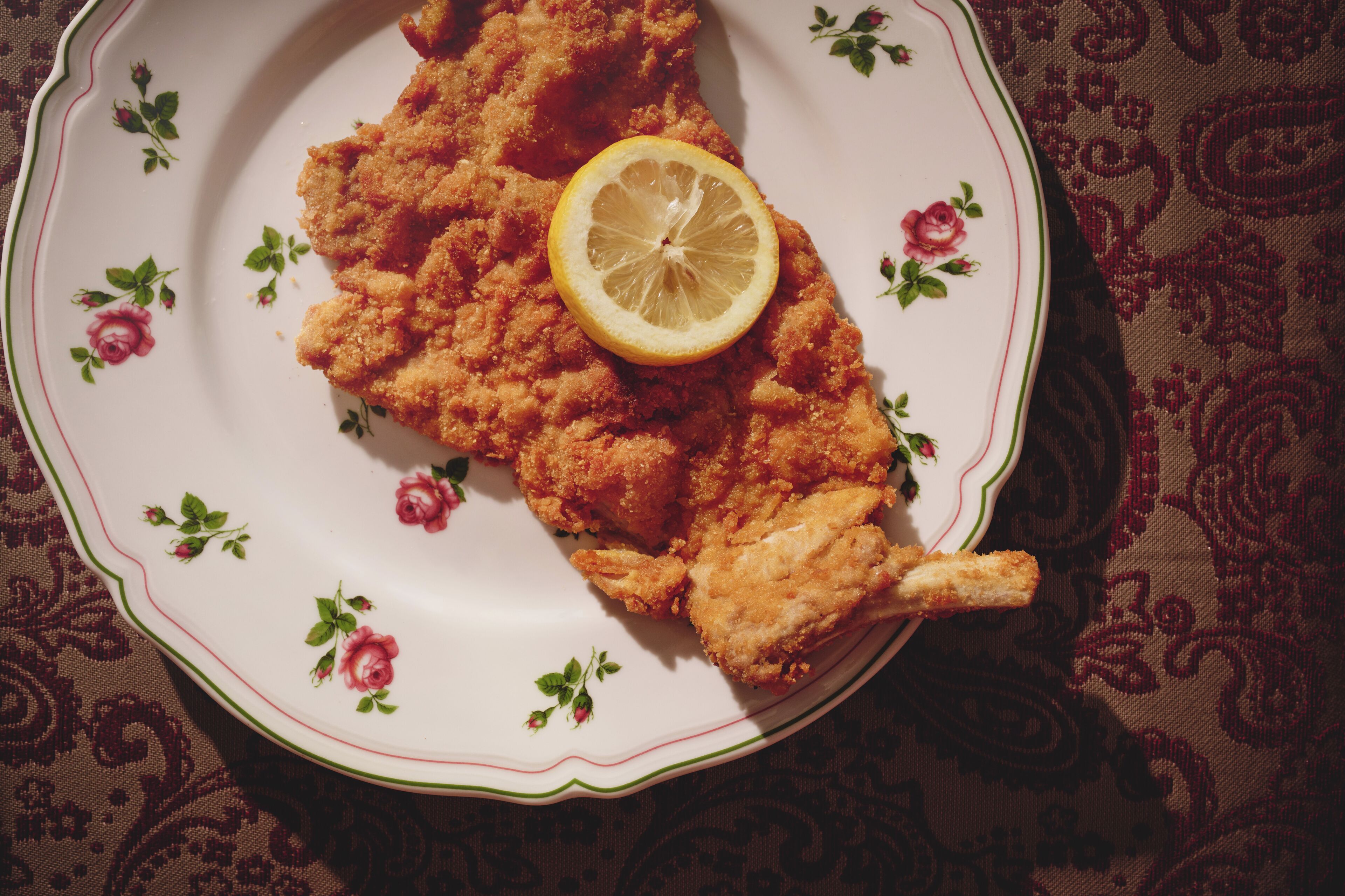 A plate with breaded chicken cutlet and a lemon slice on a floral-patterned plate.
