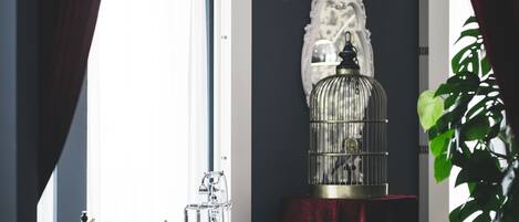 A bathroom with a freestanding bathtub, a chandelier, a birdcage, and a window with curtains.