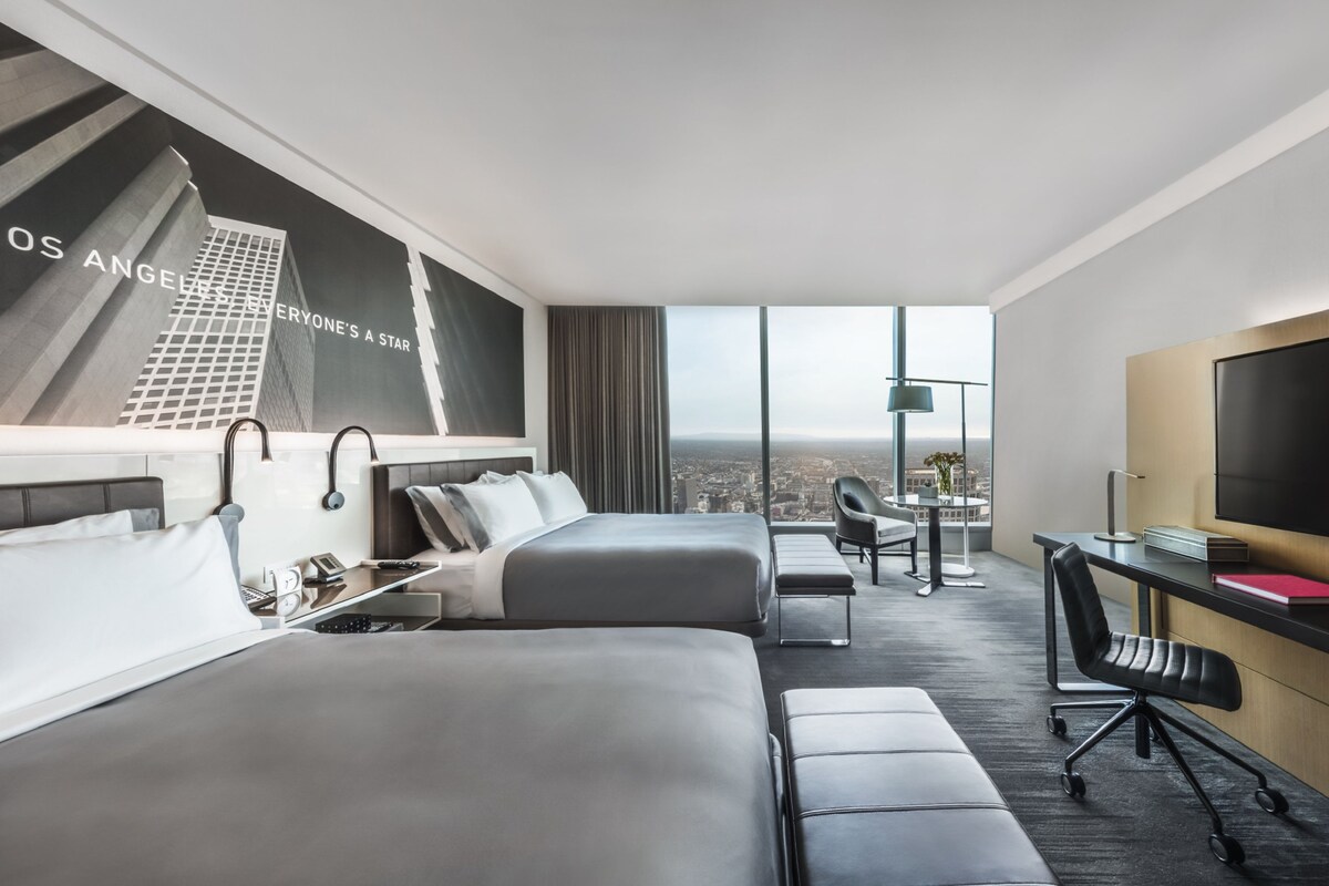 A hotel room with two beds, a desk, a chair, a TV, and a large window with a city view.